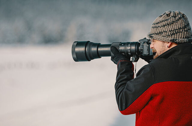 Photographer in a coat and hat shooting landscape photos using a telephoto lens