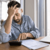 A man in a denim shirt sits at a table looking stressed while using a calculator, surrounded by bills and a laptop in a brightly lit high-rise apartment.