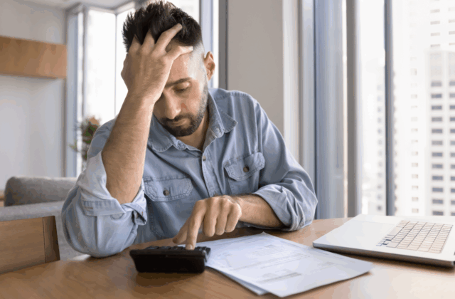 A man in a denim shirt sits at a table looking stressed while using a calculator, surrounded by bills and a laptop in a brightly lit high-rise apartment.