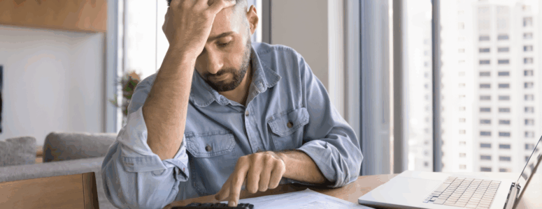 A man in a denim shirt sits at a table looking stressed while using a calculator, surrounded by bills and a laptop in a brightly lit high-rise apartment.
