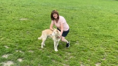 Professional pet sitter Deirdre B. kneeling in the grass with a white dog she has taken on a walk