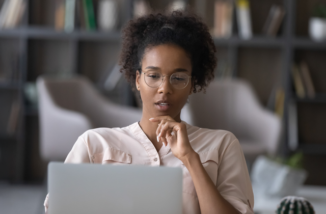 A focused young woman with glasses sits in a modern room, thoughtfully looking at her laptop screen with her hand resting near her chin.