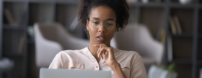 A focused young woman with glasses sits in a modern room, thoughtfully looking at her laptop screen with her hand resting near her chin.