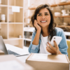 Smiling woman seated at a workspace with cardboard shipping boxes, a laptop, and a smartphone, suggesting a small business or e-commerce setting. She wears a denim shirt and appears cheerful and engaged.