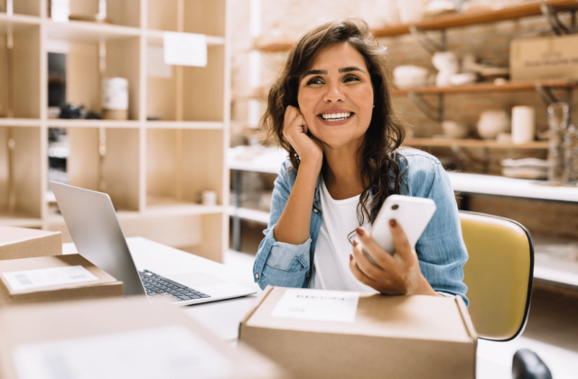 Smiling woman seated at a workspace with cardboard shipping boxes, a laptop, and a smartphone, suggesting a small business or e-commerce setting. She wears a denim shirt and appears cheerful and engaged.