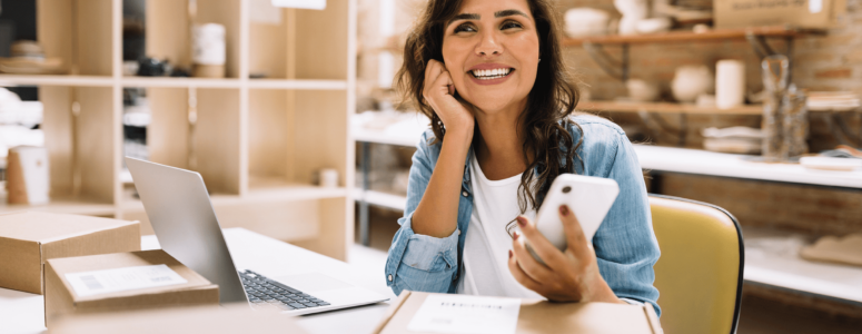 Smiling woman seated at a workspace with cardboard shipping boxes, a laptop, and a smartphone, suggesting a small business or e-commerce setting. She wears a denim shirt and appears cheerful and engaged.