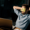 Man in a light blue shirt sits at a desk with arms behind his head and yellow sticky notes with drawn eyes covering his eyes, appearing to pretend to be alert while actually resting.