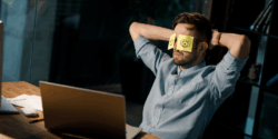 Man in a light blue shirt sits at a desk with arms behind his head and yellow sticky notes with drawn eyes covering his eyes, appearing to pretend to be alert while actually resting.