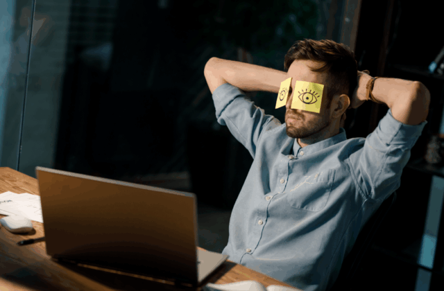 Man in a light blue shirt sits at a desk with arms behind his head and yellow sticky notes with drawn eyes covering his eyes, appearing to pretend to be alert while actually resting.