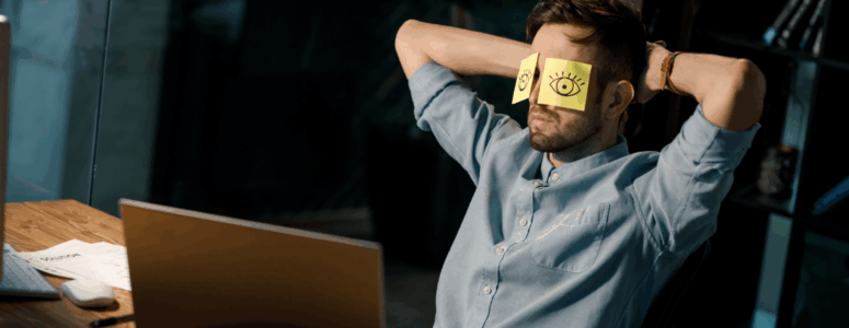 Man in a light blue shirt sits at a desk with arms behind his head and yellow sticky notes with drawn eyes covering his eyes, appearing to pretend to be alert while actually resting.
