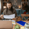 Man with long hair and glasses reviewing papers at a wooden desk with a laptop, notebooks, and coffee mug in a cozy, well-lit living room. He appears focused and serious, suggesting engagement with remote or freelance work.