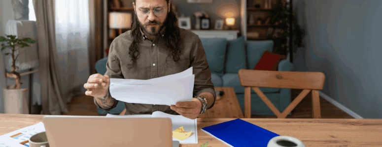Man with long hair and glasses reviewing papers at a wooden desk with a laptop, notebooks, and coffee mug in a cozy, well-lit living room. He appears focused and serious, suggesting engagement with remote or freelance work.
