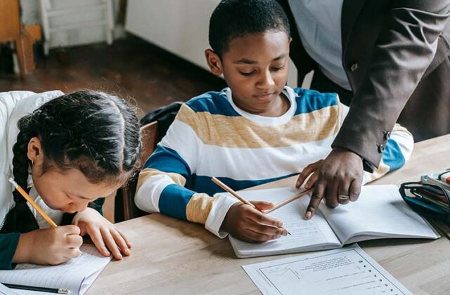 Two students sitting at a table and a teacher pointing to a student's worksheet