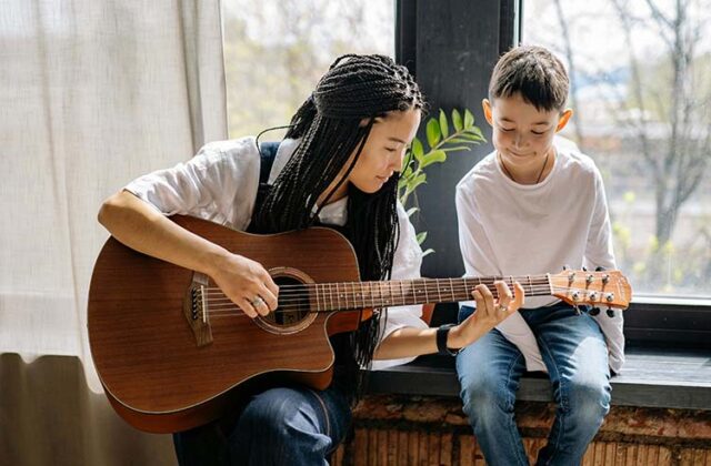 Private music teacher sitting on a bench next to a student and demonstrating guitar-playing techniques