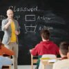Middle school teacher standing at the front of a classroom of students in front of a chalkboard displaying classwork