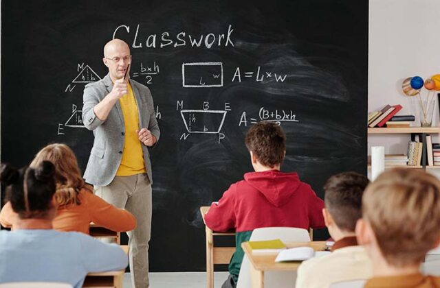 Middle school teacher standing at the front of a classroom of students in front of a chalkboard displaying classwork