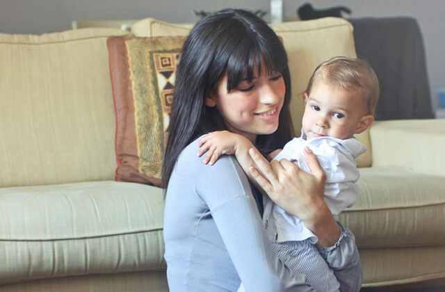 Young babysitter sitting on the floor in front of a couch holding a toddler