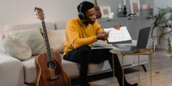 Musician sitting on a coach next to a guitar in front of an open laptop while holding a diagram