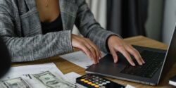 Close view of a finance professional typing on their laptop with a calculator nearby, suggesting they're working on their side hustle.