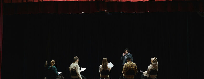 Actor standing on a stage in front of four students who are sitting and holding scripts