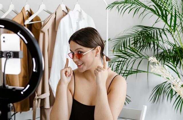 Fashion influencer sitting in front of a phone camera wearing pink sunglasses and pointing to them for viewers to see