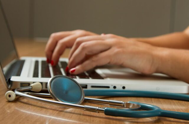 Close view of a nurse writer's hands on their laptop with a stethoscope sitting next to it.