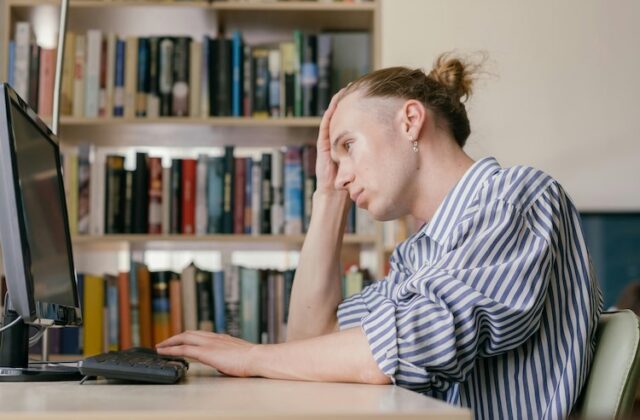A man sitting in front of a computer with one hand on his forehead, looking frustrated at the screen.