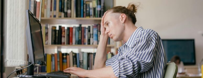 A man sitting in front of a computer with one hand on his forehead, looking frustrated at the screen.