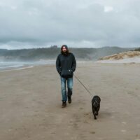 Professional dog walker wearing a hoodie and walking along the beach with a medium-sized black dog on a leash