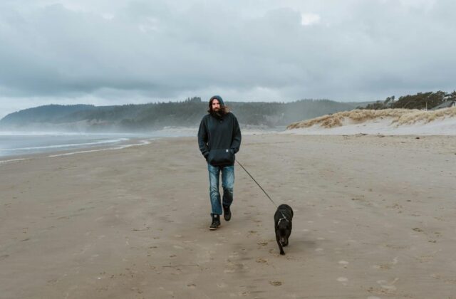 Professional dog walker wearing a hoodie and walking along the beach with a medium-sized black dog on a leash