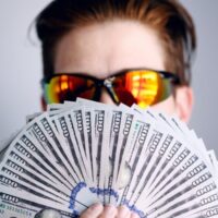 Professional influencer in sunglasses holding fanned out hundred-dollar bills in front of their face