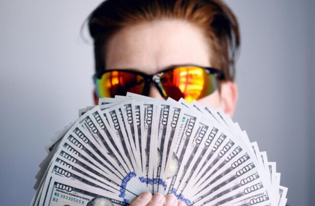 Professional influencer in sunglasses holding fanned out hundred-dollar bills in front of their face