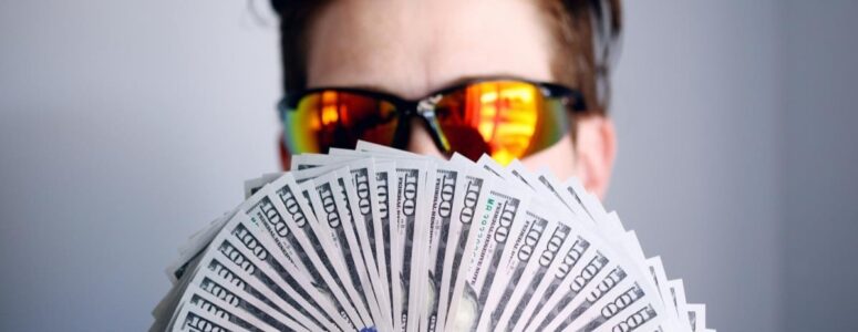 Professional influencer in sunglasses holding fanned out hundred-dollar bills in front of their face