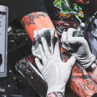 White-gloved hands of a professional tattoo artist using a tattoo needle to draw a tattoo on a client's arm