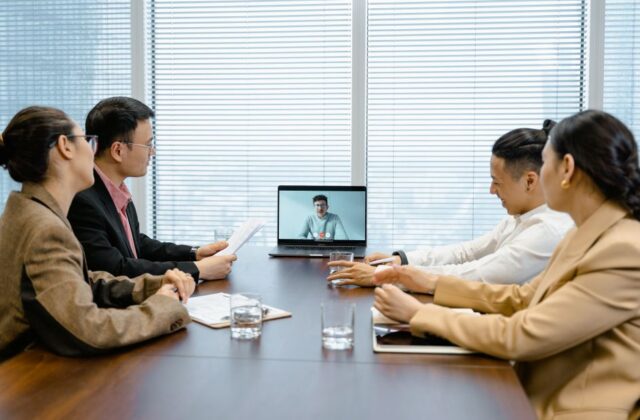 Four office-based employees gather around a table for a hybrid meeting, visually separated from their remote colleague on a laptop screen.