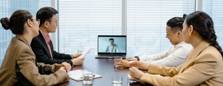 Four office-based employees gather around a table for a hybrid meeting, visually separated from their remote colleague on a laptop screen.