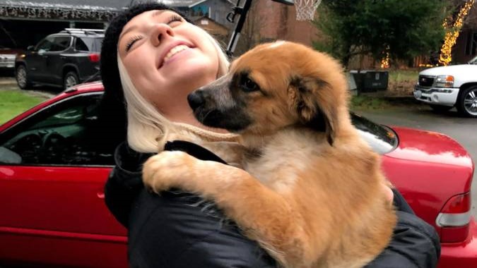 Professional pet sitter Jennifer D. smiling and holding a brown puppy in front of a red car