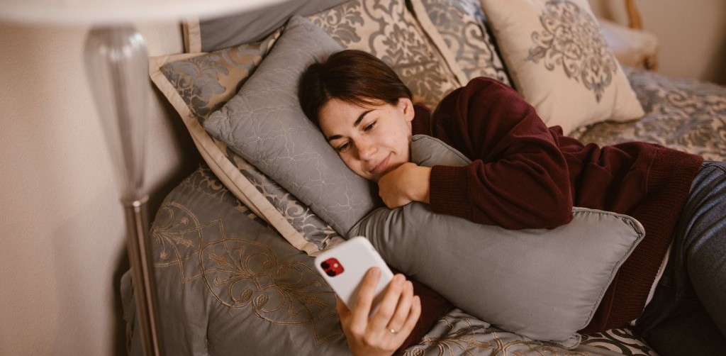 Friendly-looking girl lying in bed and smiling at messages on her phone.