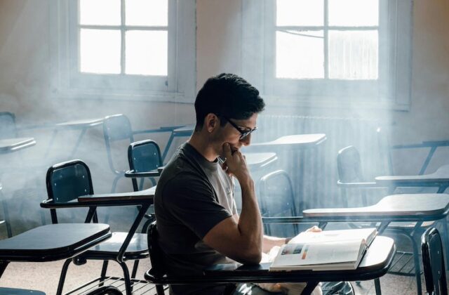 College student in empty classroom reading a book with light shining in.