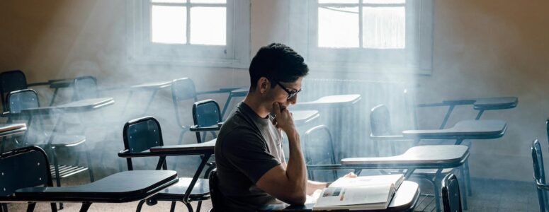 College student in empty classroom reading a book with light shining in.