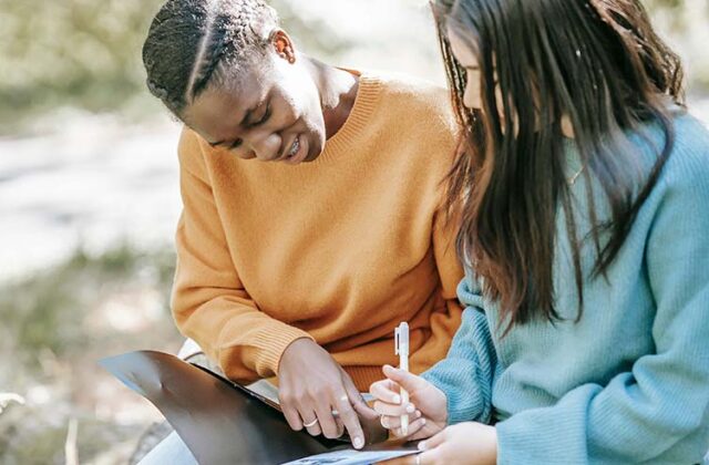 Professional tutor sitting with a student outside and guiding them through a textbook