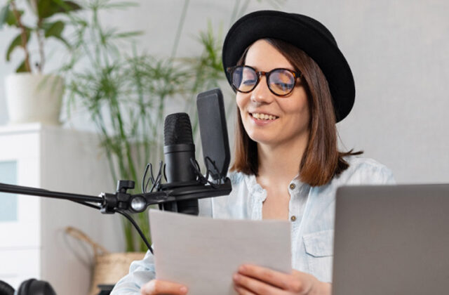 Voice actor wearing a black hat and glasses, reading from a document and speaking into a microphone