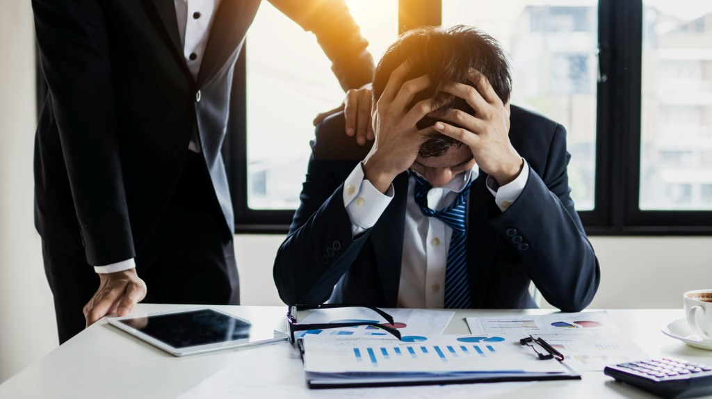 A stressed worker holding his head in his hands while his manager tries to pressure him into a promotion.